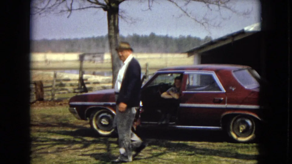 A man in a fedora, white shirt, and dark jacket walks across grass in front of a maroon sedan, with a wooden farm building, split-rail fence, open field, and tree line behind him.