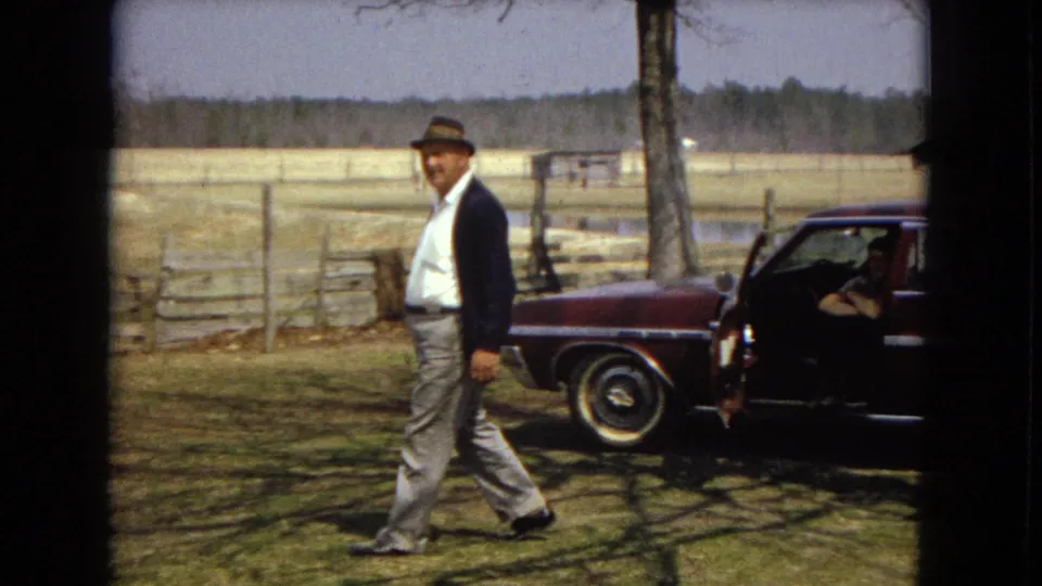 The walking man is closer to the camera, crossing a grassy farmyard while a maroon sedan with an open door and a seated adult remains in the background near fencing and open fields.