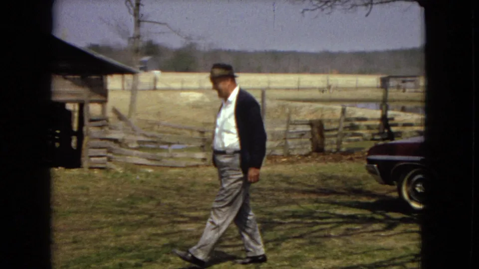 Near the end of the clip, the man in a hat and dark jacket continues walking across grass, with the maroon sedan partly cropped at the right edge and a fence, field, and farm structure behind him.
