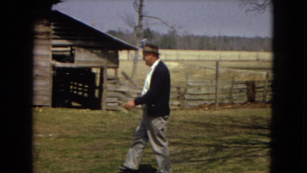 A man in a dark cardigan, light trousers, and hat walks left to right across a dry rural yard near a weathered wooden barn, consistent with the supplied 1967 United States home-movie metadata.