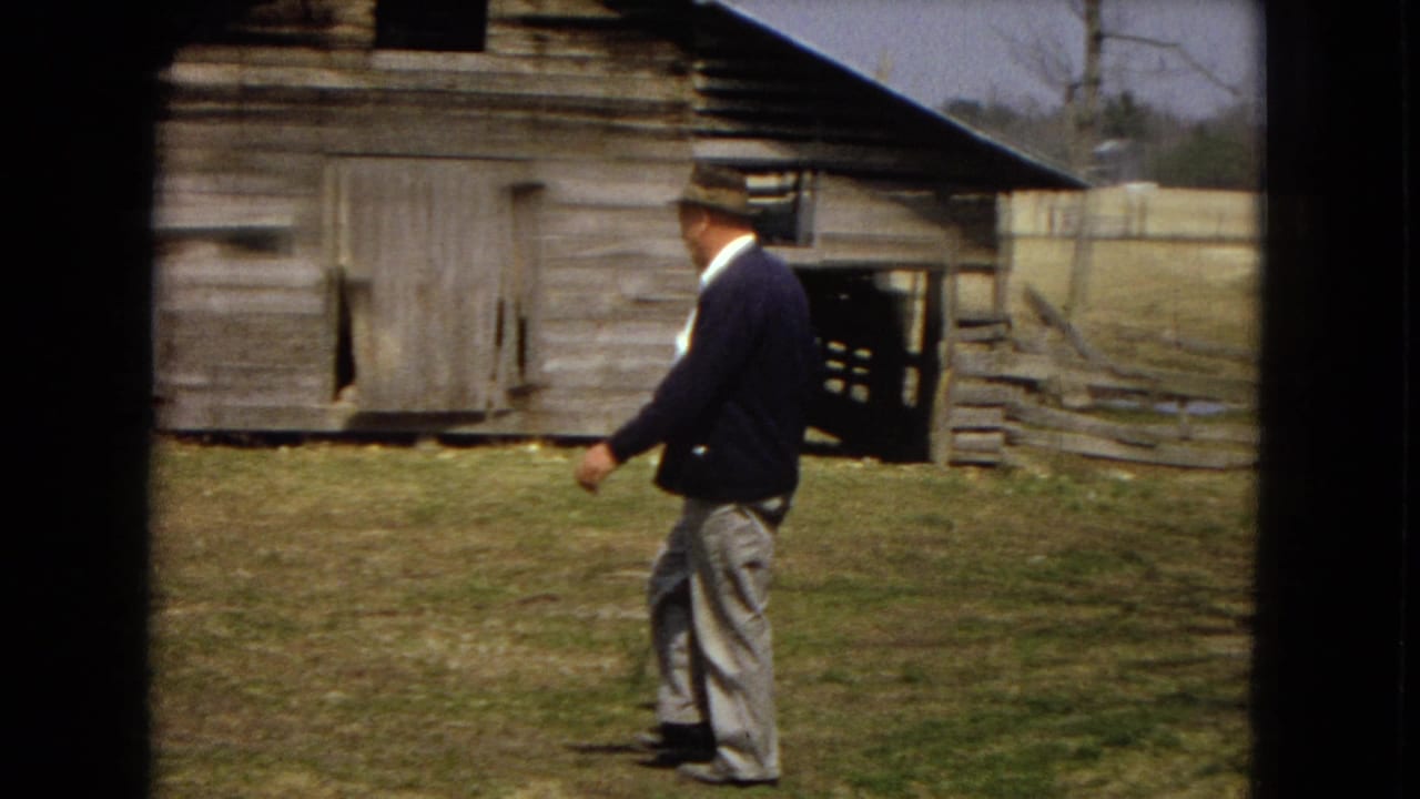 Man stands or walks near a weathered barn with fencing behind him.