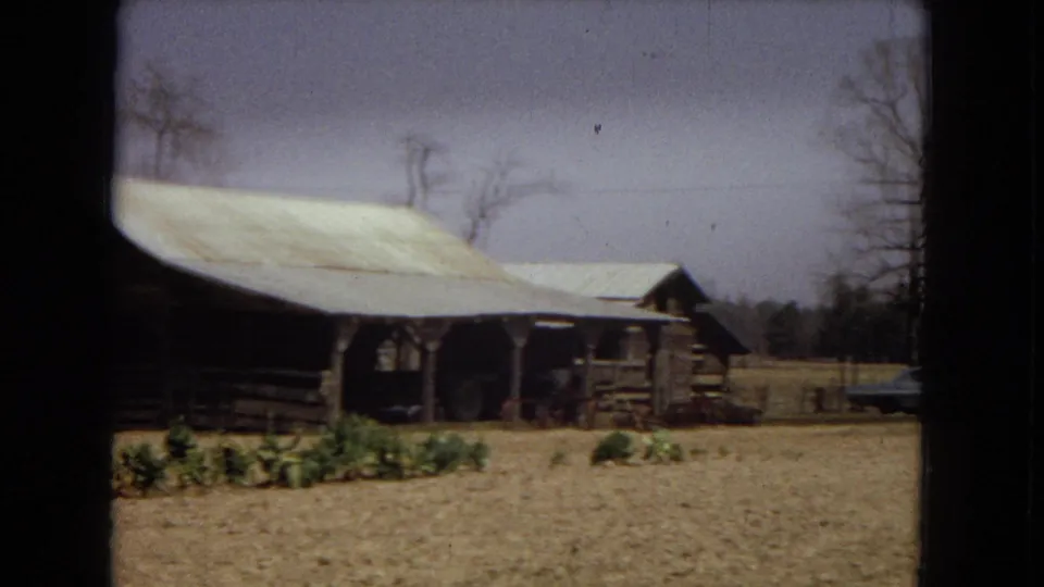A second view of the rustic farmhouse shows the pale roof, porch area, wood fencing, bare trees, dry ground, and distant rural structures under soft 8mm color.