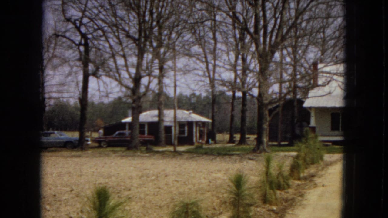 1967 U.S. 8mm frame showing a modest dark bungalow with a front porch, cars parked beside it, bare trees, and open yard space.