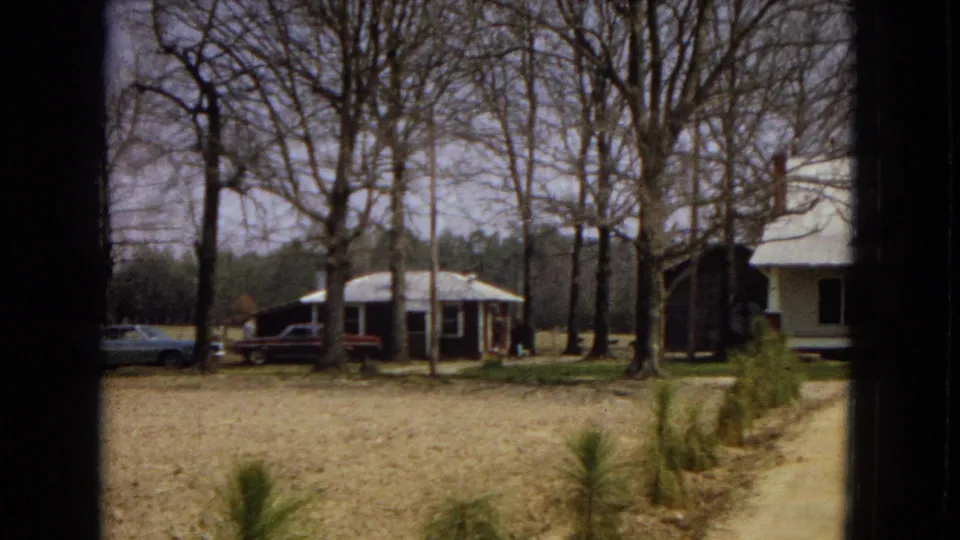 1967 U.S. 8mm frame showing a modest dark bungalow with a front porch, cars parked beside it, bare trees, and open yard space.