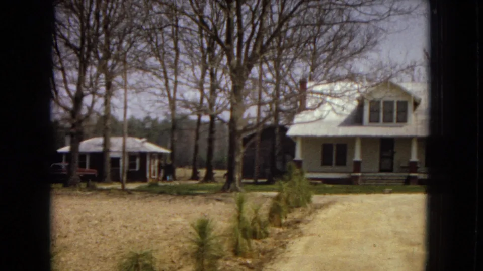 1967 U.S. home-movie frame during a pan across a rural property, with a dark bungalow at left, a white house at right, leafless trees, and a dirt driveway.