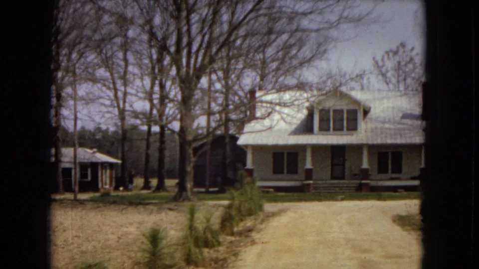 1967 U.S. 8mm frame showing a larger white house with a front porch, red-toned columns, young pine saplings along a dirt drive, and a smaller dark building nearby.