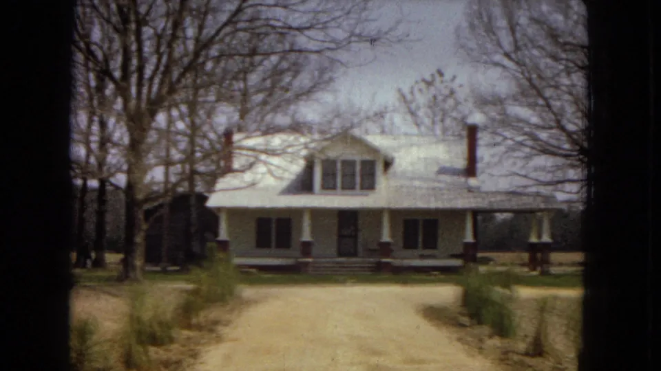 1967 U.S. home-movie frame centered on a white farmhouse-style residence with a front porch, chimney, dirt driveway, young saplings, and leafless trees around the yard.