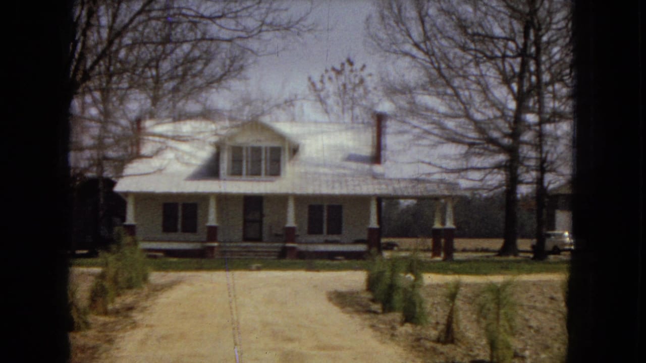 1967 home-movie frame showing a white rural farmhouse with a broad porch, red brick column bases, a dirt driveway, young evergreens, and leafless trees.