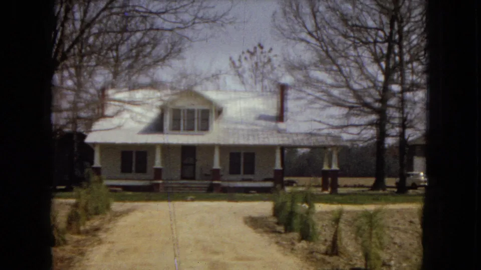 1967 home-movie frame showing a white rural farmhouse with a broad porch, red brick column bases, a dirt driveway, young evergreens, and leafless trees.