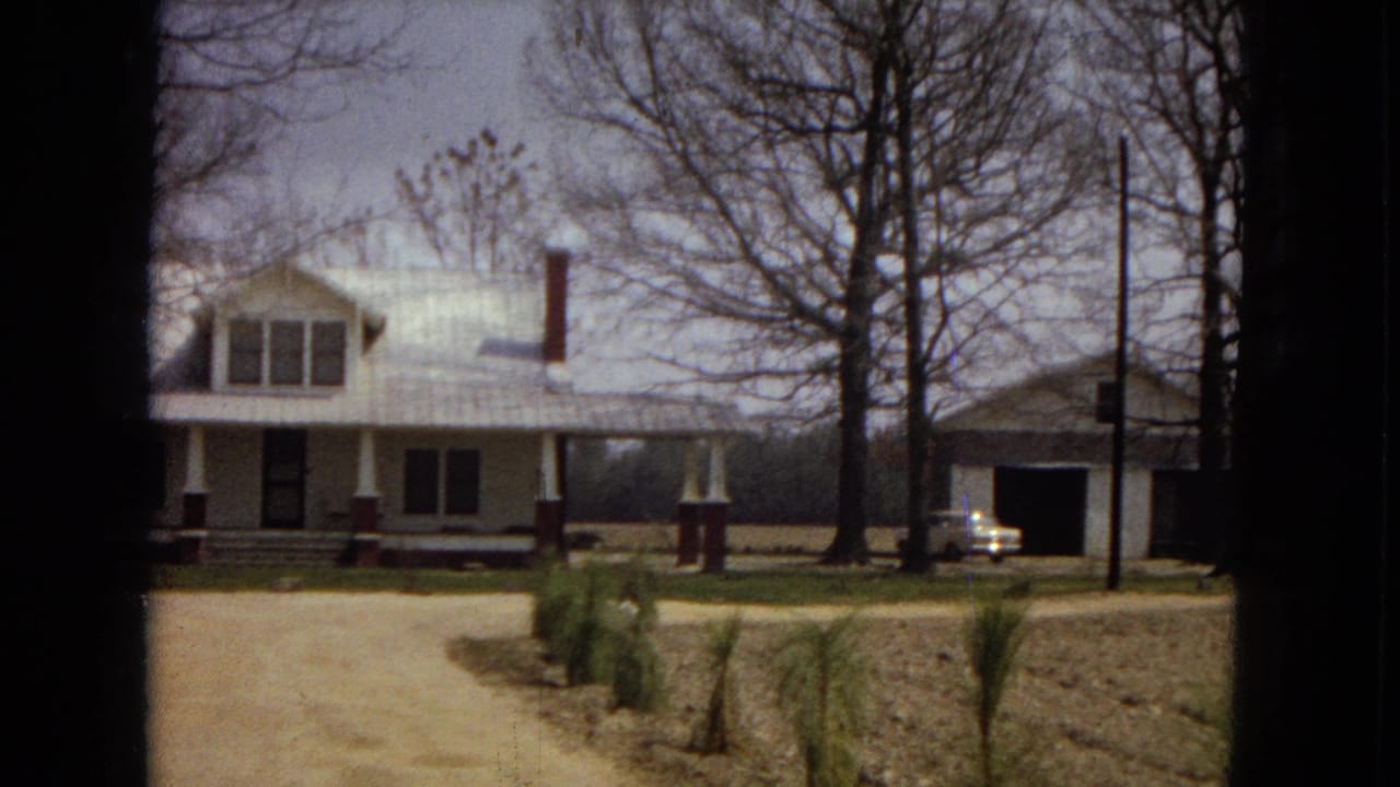The pan holds on the farmhouse and dirt drive, with an older light-colored car parked near a dark garage or outbuilding at the right edge.