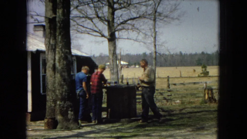 Three men in casual clothing stand near a tree and table beside a rural building, with open fields and a fence visible in the background.