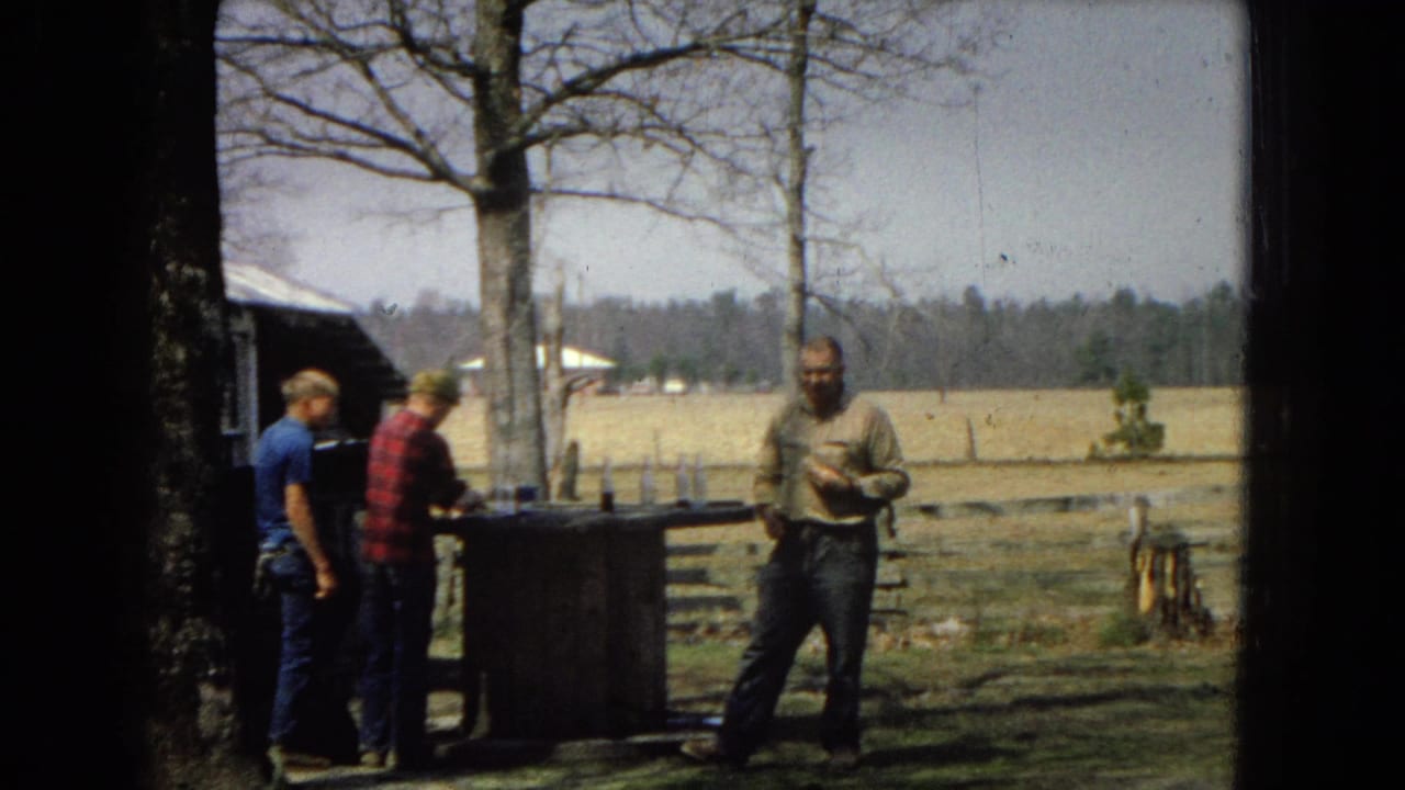 In a 1967 United States 8mm home-movie frame, three adult men gather outdoors near a wooden work table beside trees, fencing, and open fields.