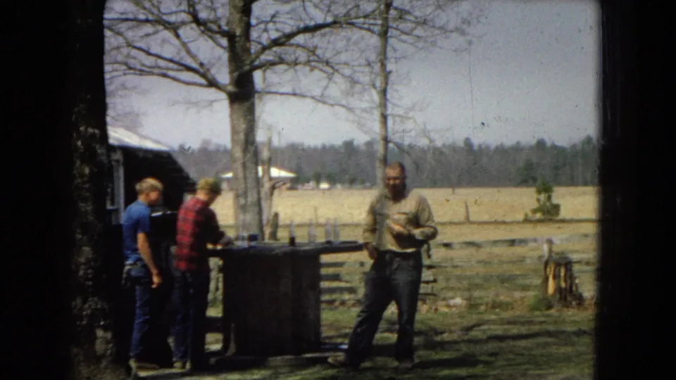 In a 1967 United States 8mm home-movie frame, three adult men gather outdoors near a wooden work table beside trees, fencing, and open fields.