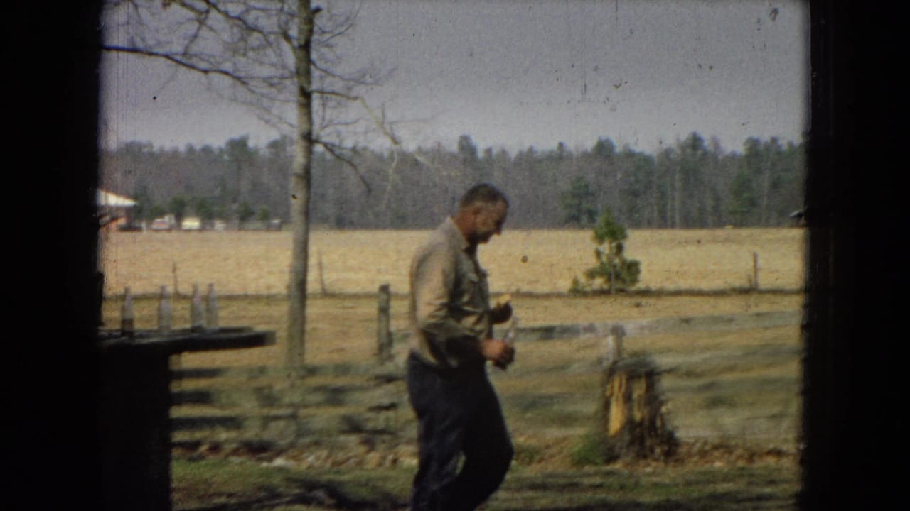 An older man walks across a fenced yard holding a small object.