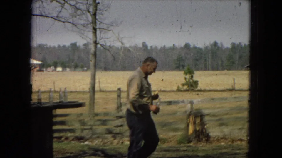 A man in a light shirt walks through a rural fenced yard holding a small object, with open fields and distant trees behind him.