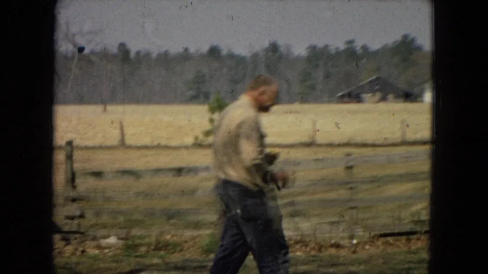 The man continues walking through the rural yard, slightly soft in motion, with fencing, a stump, open field, and a distant farm building visible.