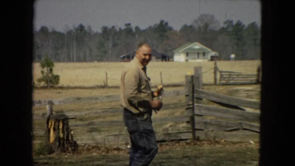 A man holding a small object pauses near a wooden fence in a rural yard, with a tree stump, open field, and distant house-like structure behind him.