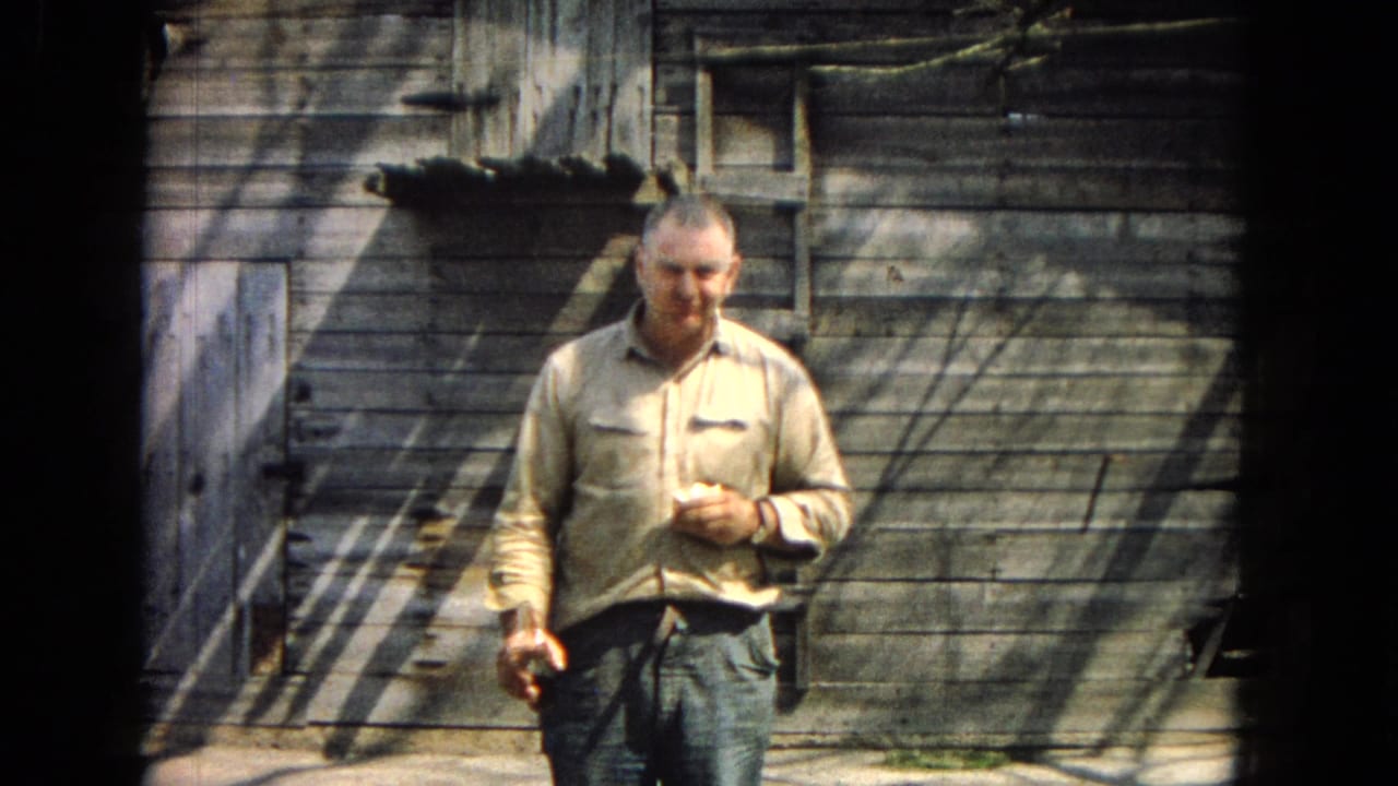 In 1967 archival 8mm footage, a man in a light shirt and jeans stands before a weathered wooden shed, holding food in one hand and a bottle in the other.
