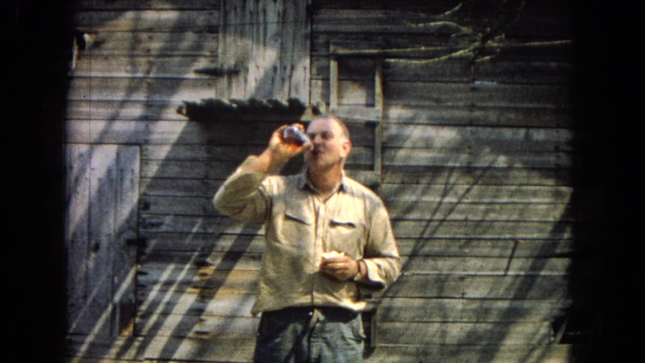 Man drinks from a bottle in front of a weathered wooden shed.