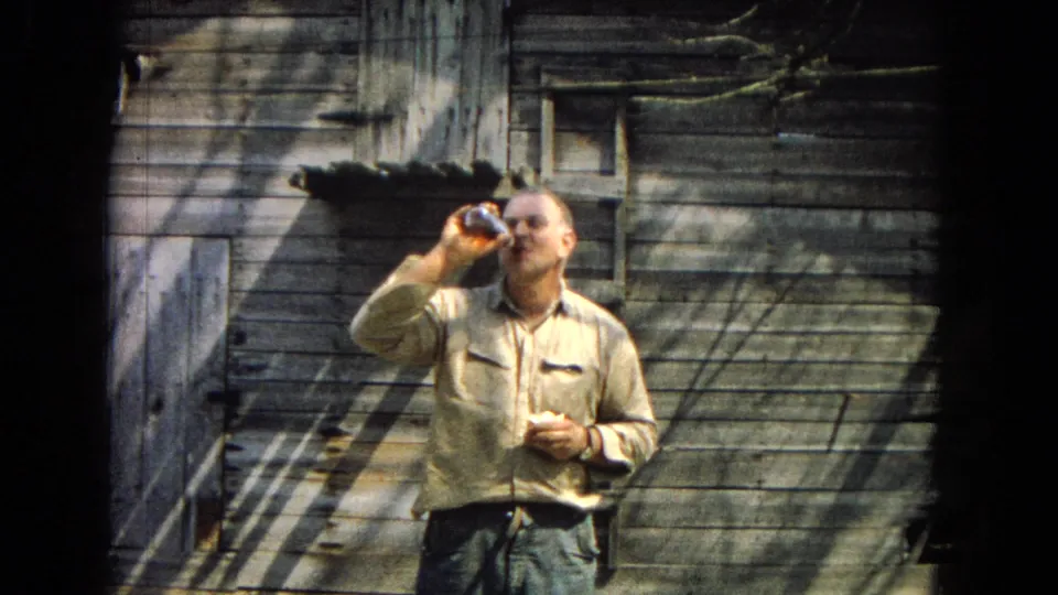 The man raises a bottle to drink while standing in front of aged wooden siding and a small shed door in this 1967 home-movie frame.