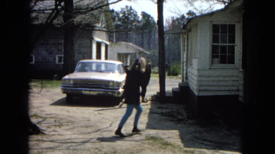 A girl in a dark coat and light pants stands on a dirt driveway near a light-colored 1960s-style car, with a white mobile home and trees behind her.