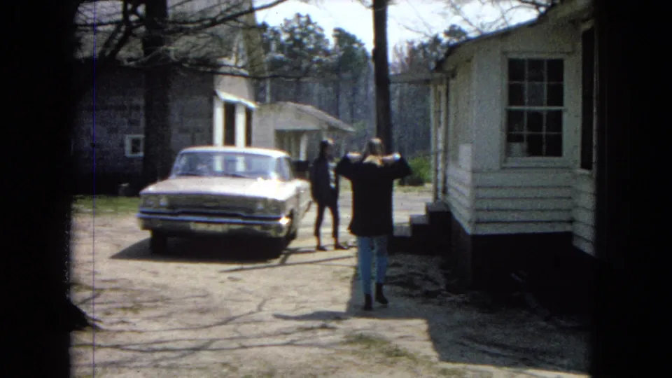 The girl stands with her back partly to the camera, facing two distant figures near a parked light-colored car beside a white mobile home and trees.