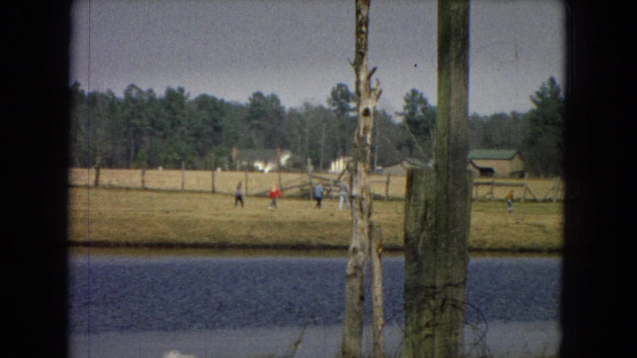 1967 8mm home-movie frame showing a distant open area near water with small figures, utility poles, trees, and dark film-edge framing.