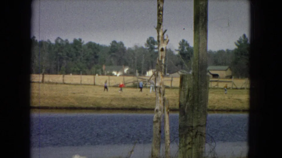 1967 8mm home-movie frame showing a distant open area near water with small figures, utility poles, trees, and dark film-edge framing.