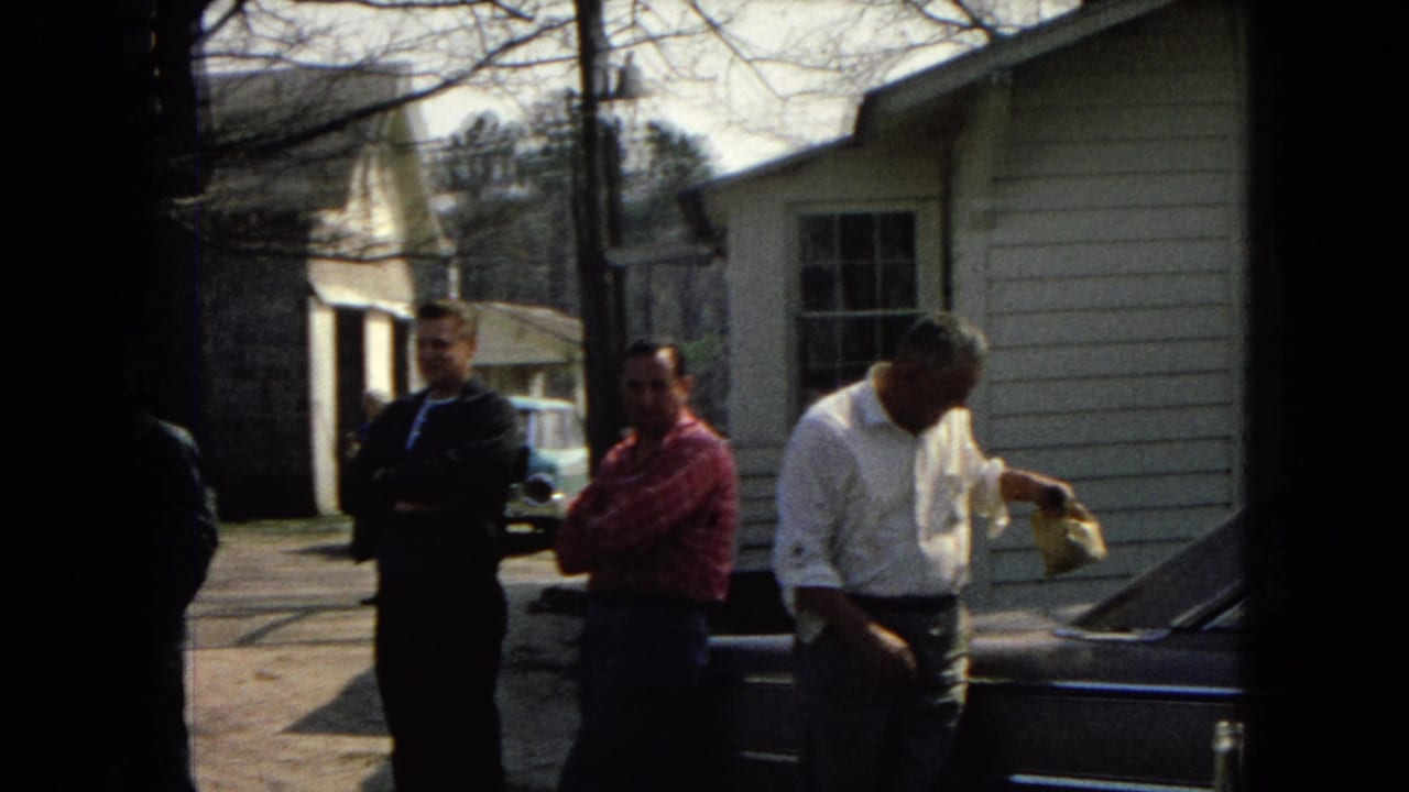 Men stand near a dark car outside a light-sided house on a sunny residential street.