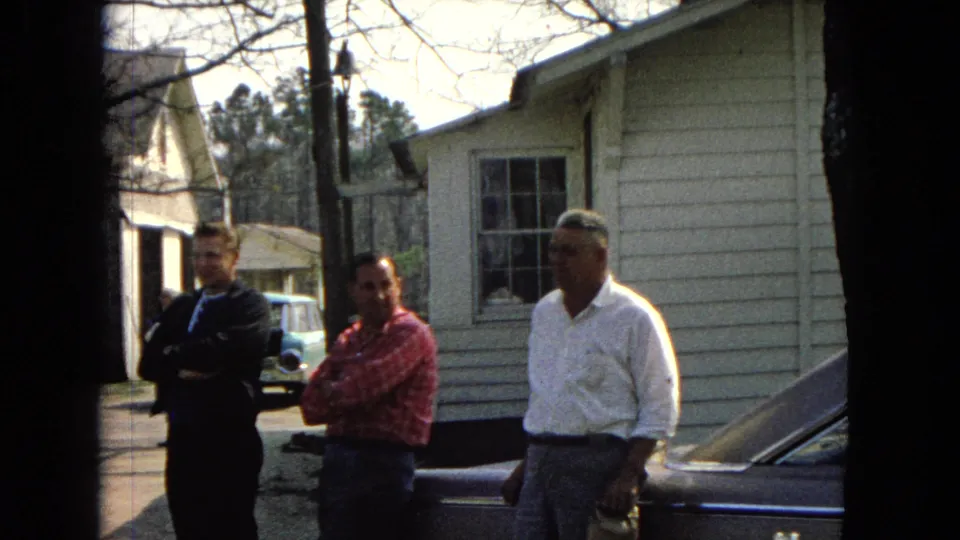 1967 United States 8mm frame of three adult men in casual dress standing by a dark car outside a modest light-sided house.