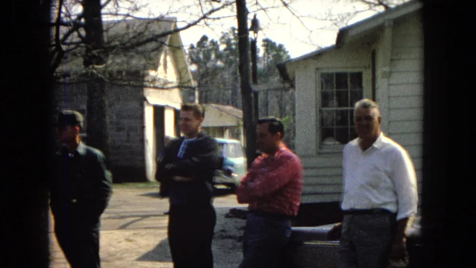 1967 United States home-movie frame showing four adult men outside beside a parked car, a light-sided house, bare branches, and nearby buildings.