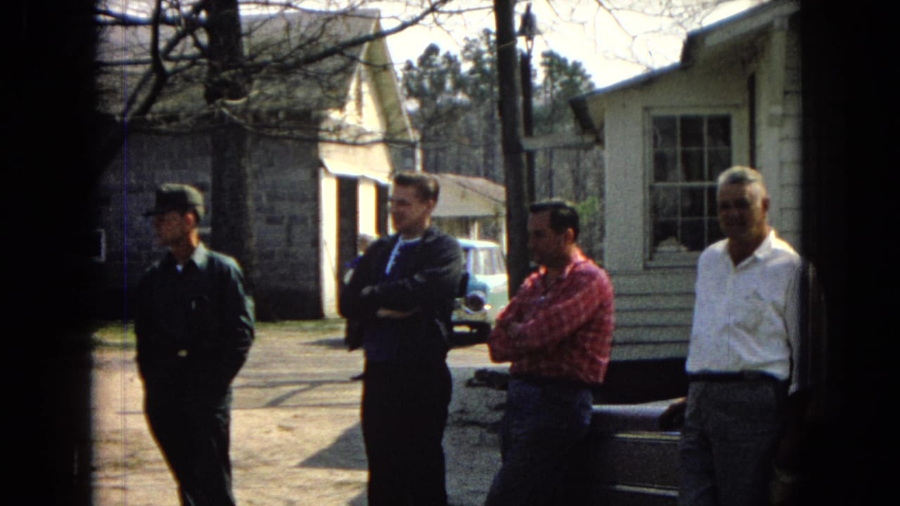 Four adult men in 1960s attire stand outdoors in a United States residential yard, with a uniformed man at left, a red plaid shirt, a white shirt, bare tree branches, small buildings, and a light blue car visible behind them.
