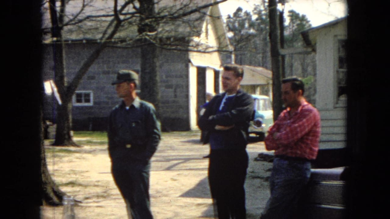 Three men stand near a house and stone building with a car partly visible behind them.