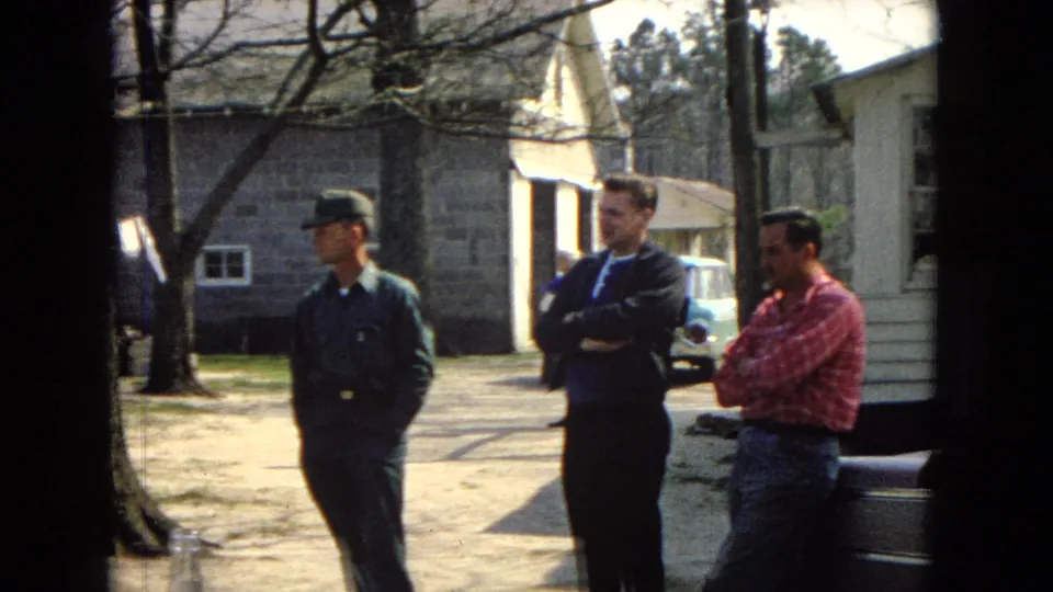 Three adult men are visible in a sunlit yard, including a man in a green uniform-style outfit and cap, a man in a dark jacket with arms crossed, and a man in a red plaid shirt near residential buildings and a partly visible car.