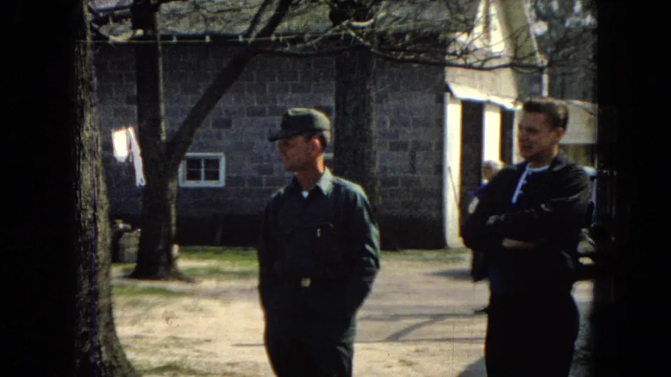 A uniformed adult man in a green cap stands beside a man in a dark jacket with crossed arms, with a stone building, window, tree trunk, dirt path, and sunlit yard visible in the background.