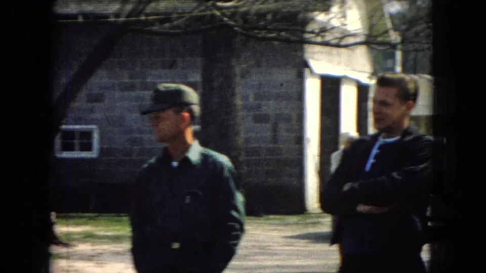Two adult men stand outdoors in front of a stone building, with one wearing a green uniform-style cap and shirt and the other wearing a dark jacket, while a doorway, small window, tree shadows, and sunlit ground appear behind them.