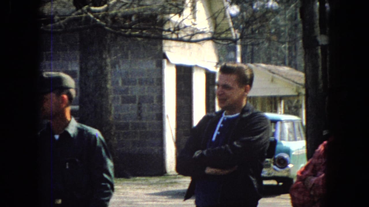 Adult men in a 1967 United States home-movie frame stand near a brick building and a light blue mid-century car, with one man in a dark cap partly visible at left.