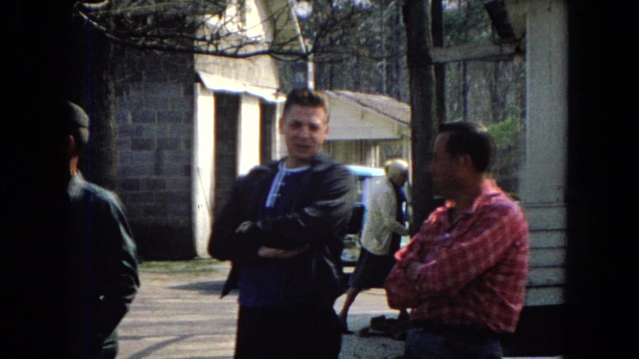 Two adult men appear to converse or wait near a white-sided structure and light blue car in a 1967 United States street-corner home-movie scene.