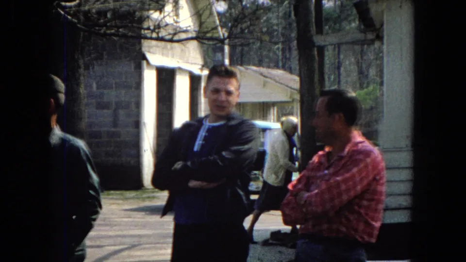 Two adult men appear to converse or wait near a white-sided structure and light blue car in a 1967 United States street-corner home-movie scene.