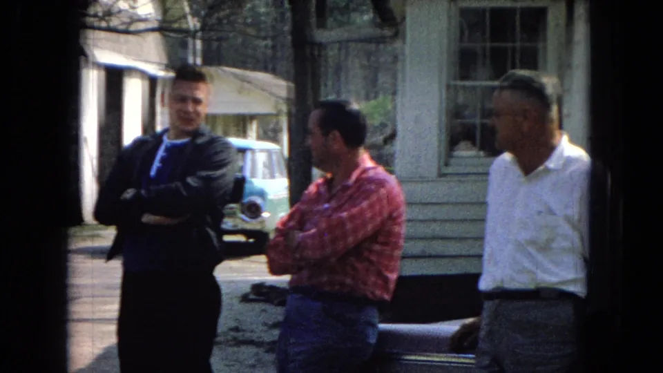 Three adult men stand casually outside a small building, including one in a red plaid shirt and one in a white button-down, with a parked car partly visible behind them.