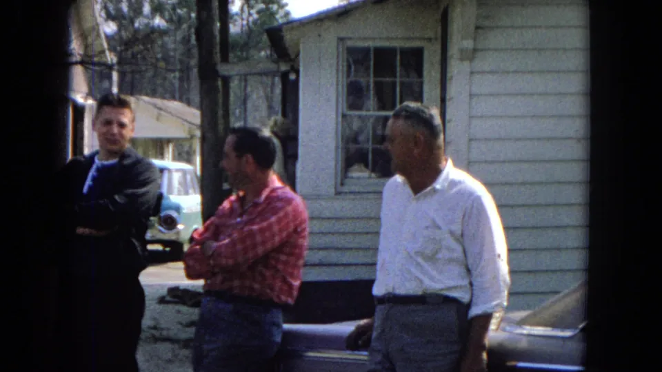 Three adult men remain in casual conversation beside a white-sided building in this 1967 United States home-movie frame, with a light blue car and window behind them.