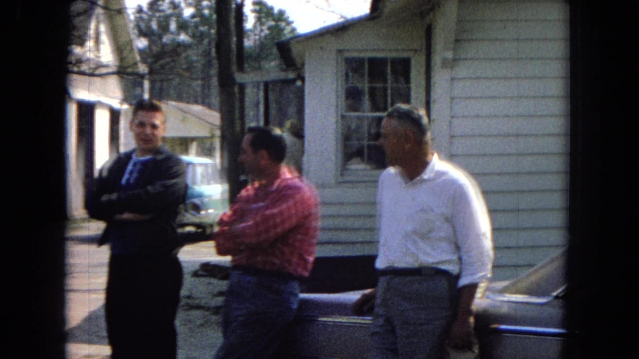 Three adult men in 1960s clothing stand outside a white clapboard building with a turquoise car partly visible nearby.