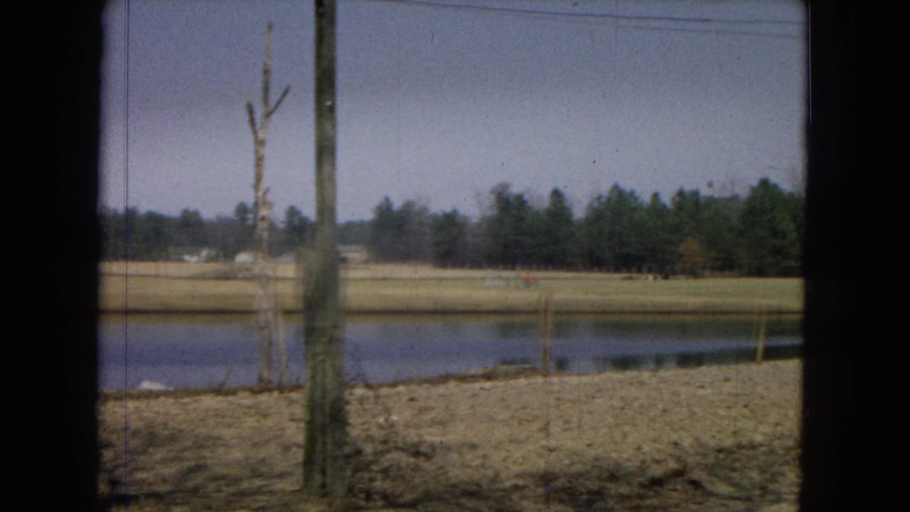 1967 8mm home-movie view in the United States showing a pond, dry rural field, leafless tree, and distant evergreen line framed by dark foreground edges.