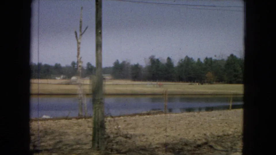 1967 8mm home-movie view in the United States showing a pond, dry rural field, leafless tree, and distant evergreen line framed by dark foreground edges.