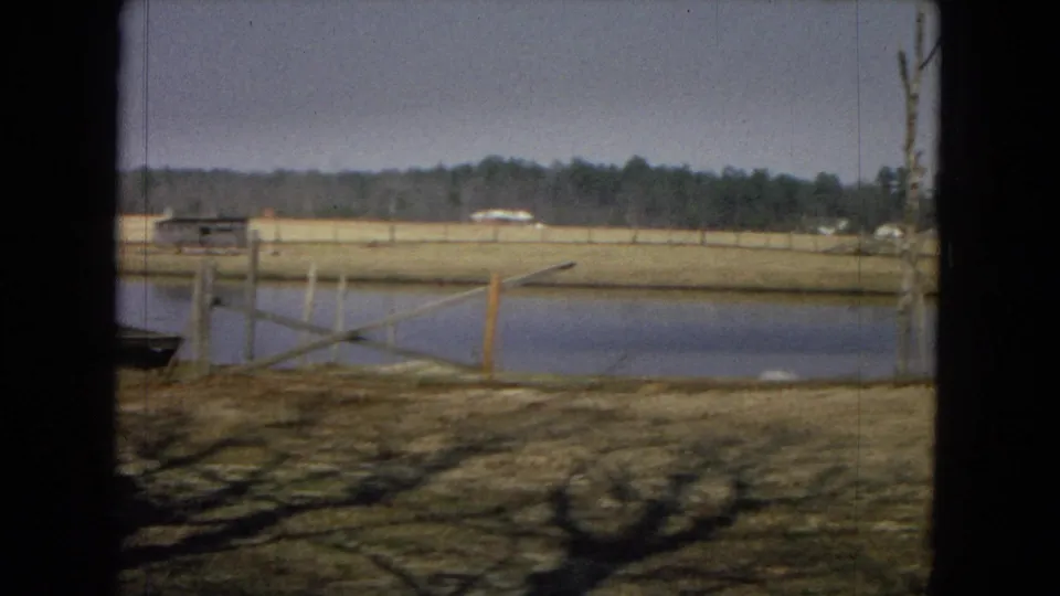1967 8mm rural United States view showing the pond edge, angled wooden rail, dry field, distant trees, and small light-colored buildings in the background.