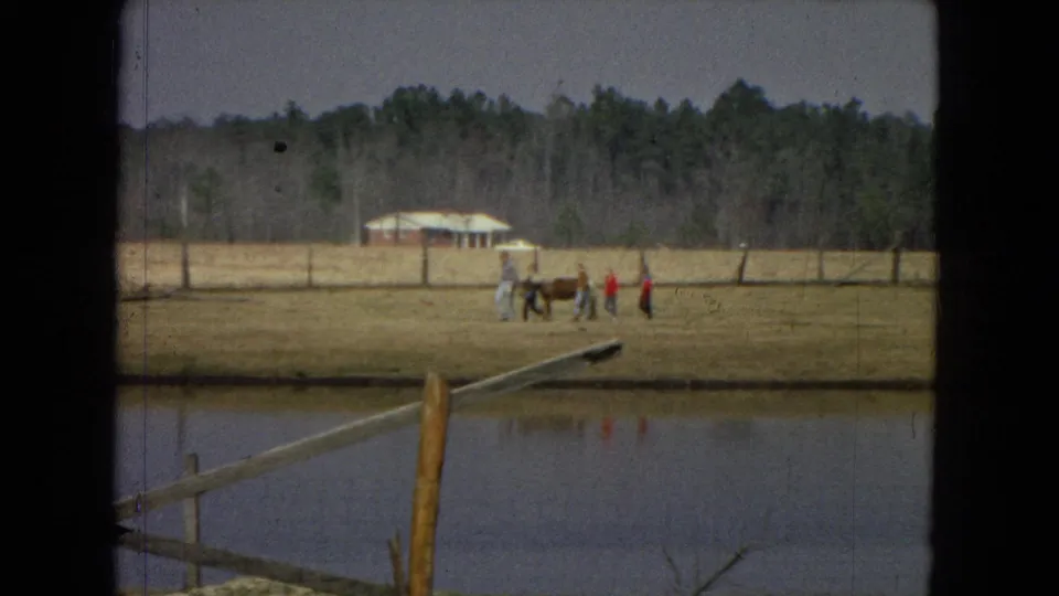 1967 United States home-movie frame showing a small group of people walking across a dry field near a pond, wooden fencing, a small rural building, and distant trees.