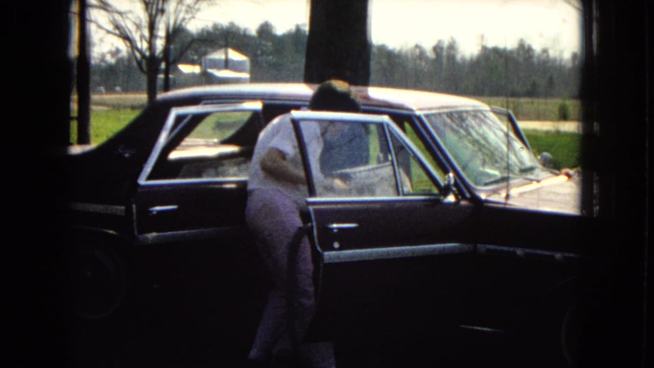In 1967 home-movie footage from the United States, a man in light clothing leans into the open rear passenger door of a dark sedan near bare trees and a rural yard.
