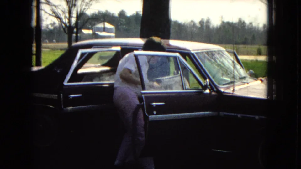 In 1967 home-movie footage from the United States, a man in light clothing leans into the open rear passenger door of a dark sedan near bare trees and a rural yard.