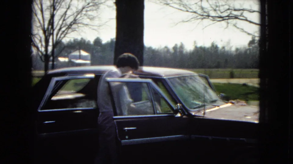 The man bends toward the sedan interior through the open rear passenger door as the camera looks out from a dark interior toward a yard and tree line.