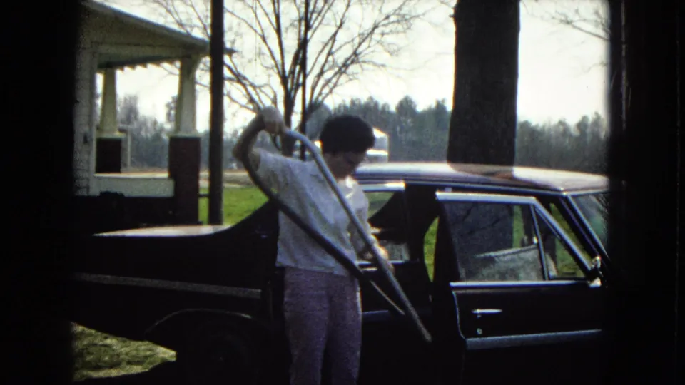 The man holds a long vacuum hose or wand beside the dark sedan, with the open rear door, porch post, bare trees, and rural yard visible around him.